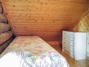 Une petite chambre avec un lit simple, un plafond en bois à pente, une commode blanche avec des tiroirs et une couvre-lit à motifs floraux.