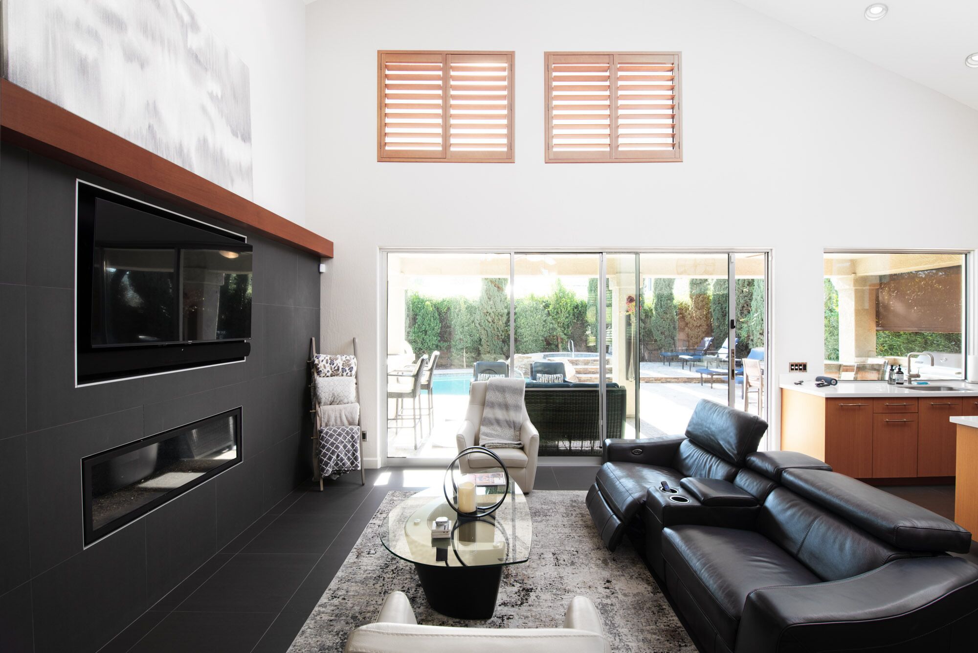 A modern living room with a black leather sofa, a glass coffee table, and a TV mounted on a dark wall. There are wooden blinds on the windows, and a sliding glass door leads to an outdoor area with a pool and seating.