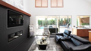 Een moderne woonkamer met een leren zwarte bank, een glazen salontafel en een televisie die aan een donkere muur is bevestigd. Er zijn houten zonwering op de ramen, en een schuifdeur van glas leidt naar een buitenruimte met een zwembad en zitplaatsen.
