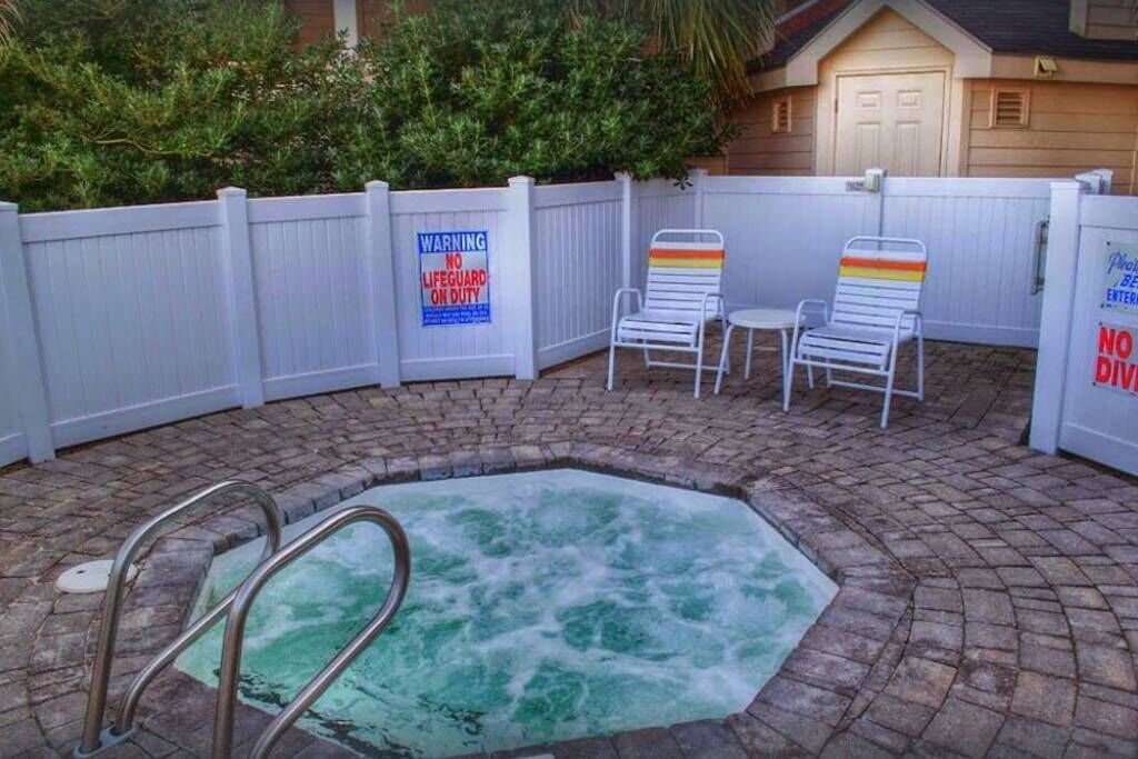 A hot tub with a wooden deck and a white fence. There are two lounge chairs and a sign warning of no lifeguard on duty.