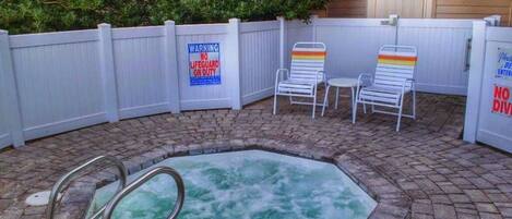 A hot tub with a wooden deck and a white fence. There are two lounge chairs and a sign warning of no lifeguard on duty.