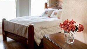 Ein ordentlich bezogenes Bett mit weißer Tagesdecke und Kissen, ein Nachttisch aus Holz mit einer Vase voller rosa Blumen und ein Fenster mit Jalousien in einem Zimmer mit Holzboden und Backsteinwand.