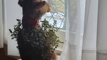 Un ours en peluche avec un nœud rouge, assis sur une surface en bois près d’une fenêtre ornée de rideaux légers.