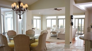 A spacious living room with a glass dining table surrounded by wicker chairs, a ceiling fan, and large windows offering a view of the outdoors.