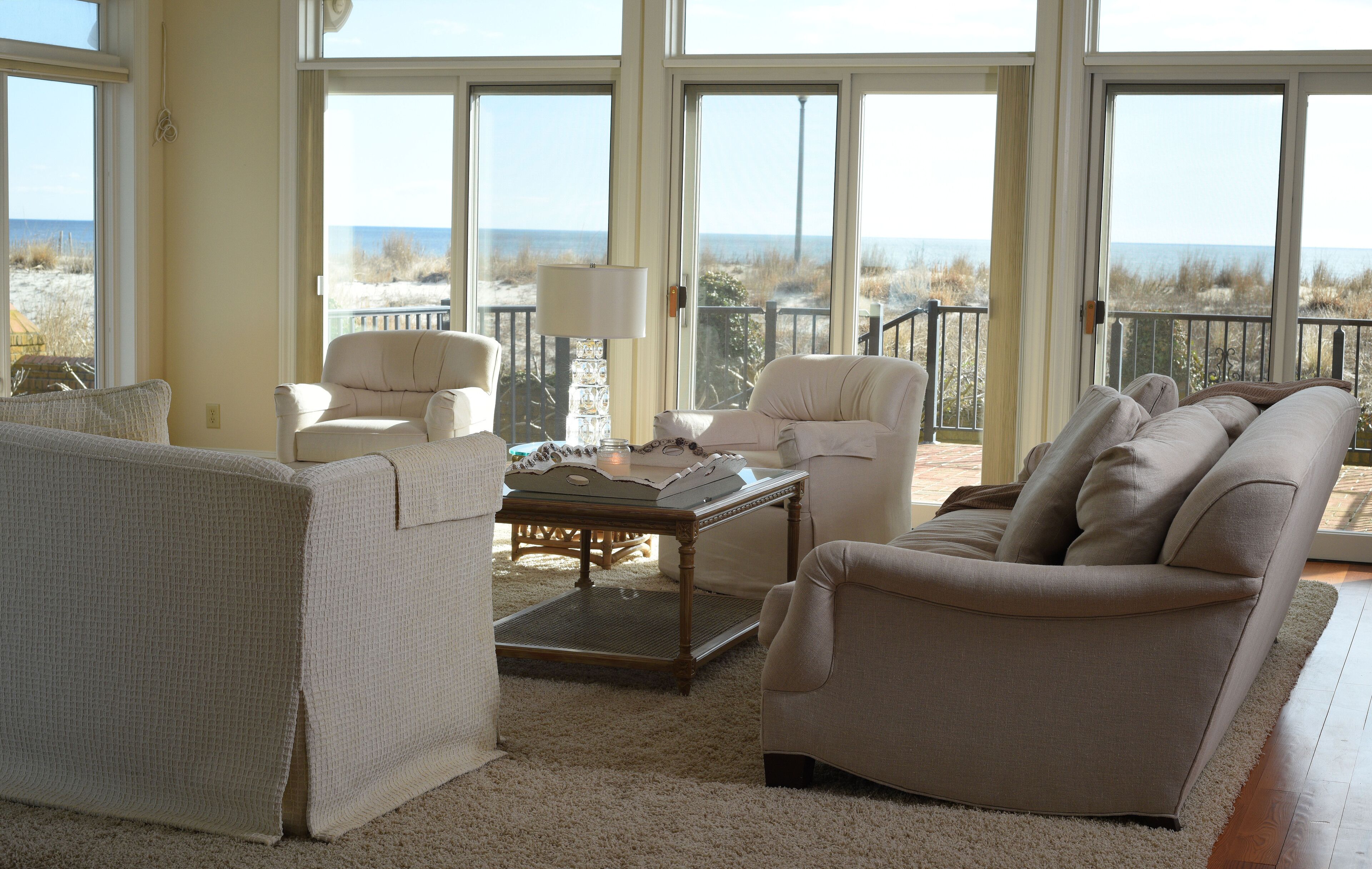 A spacious living room with large windows offering a view of the beach and ocean. There are two sofas, a coffee table, and a lamp.