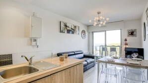 A modern kitchen with a sink, wooden cabinets, a dining area with a table and chairs, a sofa, and a TV. - Apartment Claire by Interhome (Arcachon)