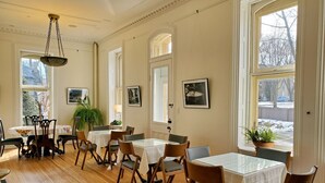 A dining area with wooden flooring, white tables, and chairs. There are large windows with views of the outside. - Governor's Mansion Inn (Madison)
