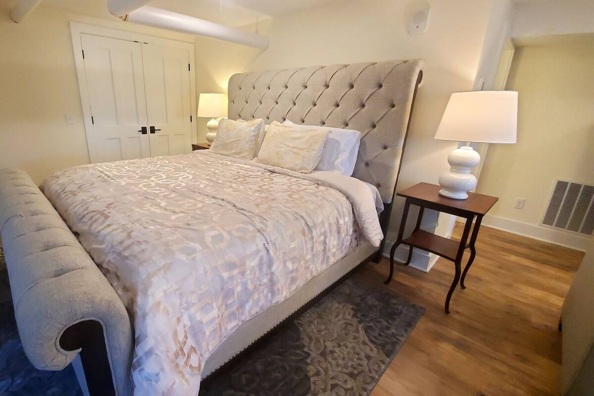A bedroom with a large bed, a bedside table with a lamp, a chair, and a door.