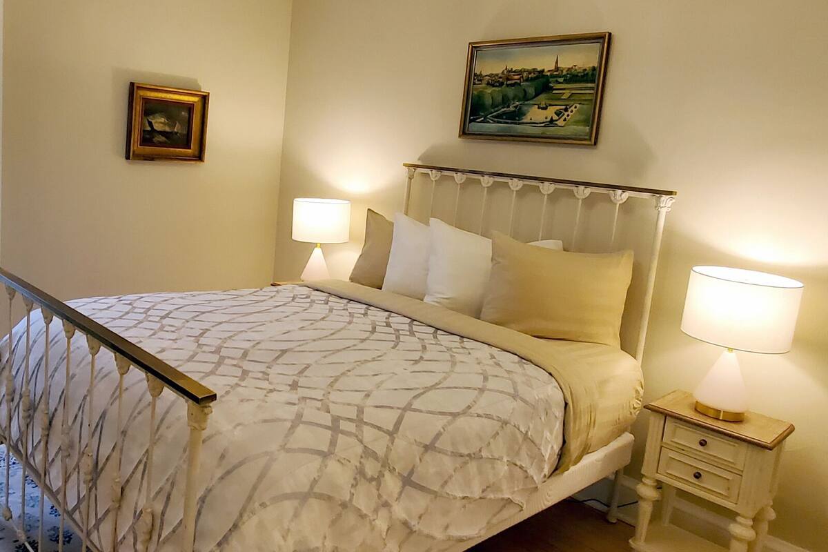 A neatly made bed with a patterned bedspread, two bedside lamps, a small bedside table with a lamp, and a framed picture on the wall.