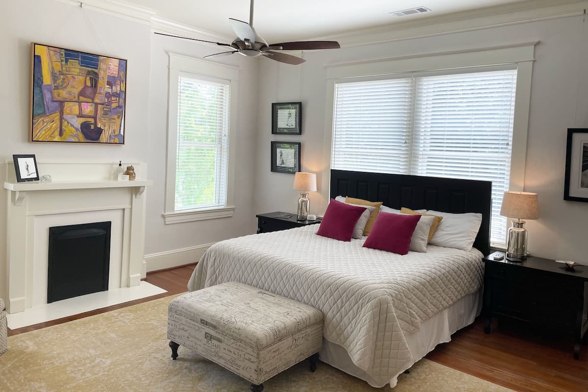 Une chambre à coucher avec un grand lit, une cheminée, un ventilateur de plafond et une fenêtre avec des stores.
