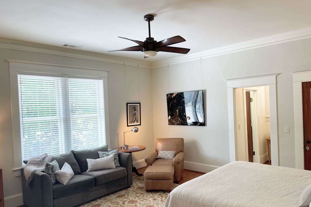 Une chambre à coucher avec un lit, un canapé gris, un fauteuil marron, un ventilateur de plafond et un tableau au mur.