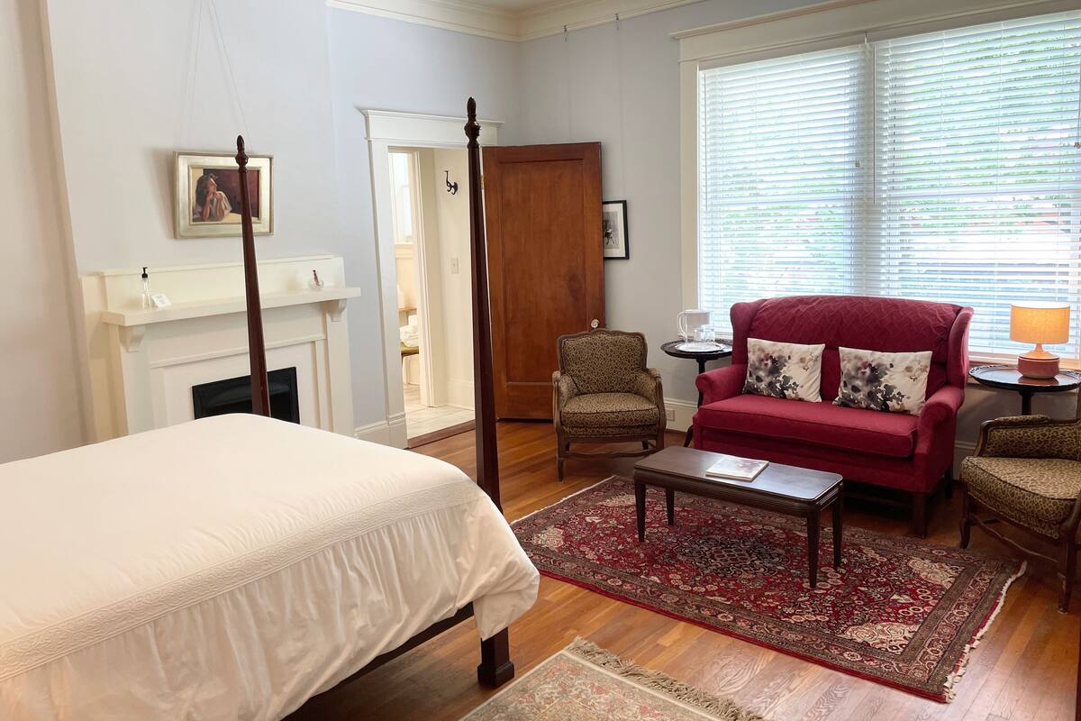 Une chambre à coucher avec un lit, un fauteuil rouge, une chaise en bois, une petite table et une cheminée.