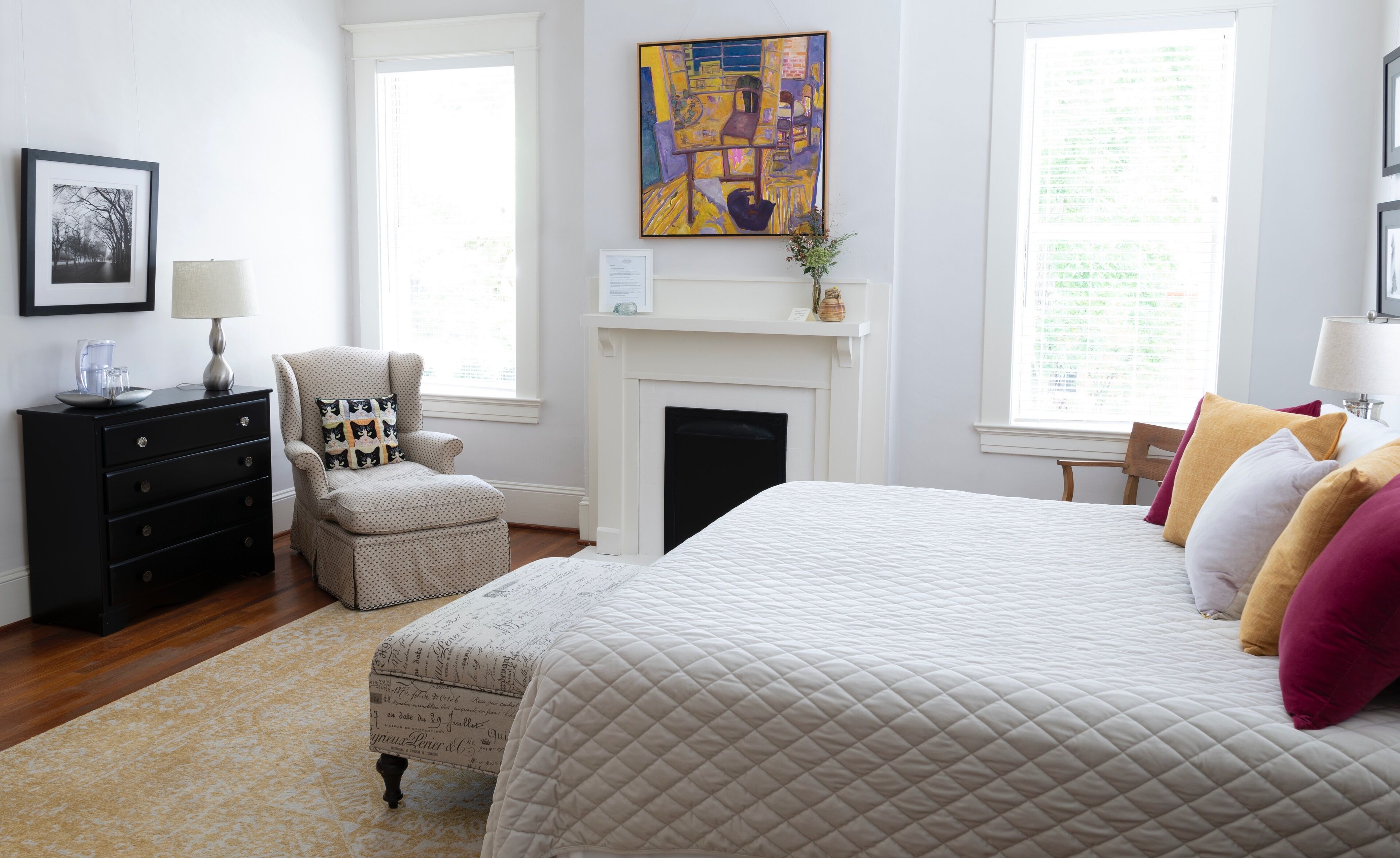 A bedroom with a bed, a chair, a dresser, a fireplace, and artwork on the wall.