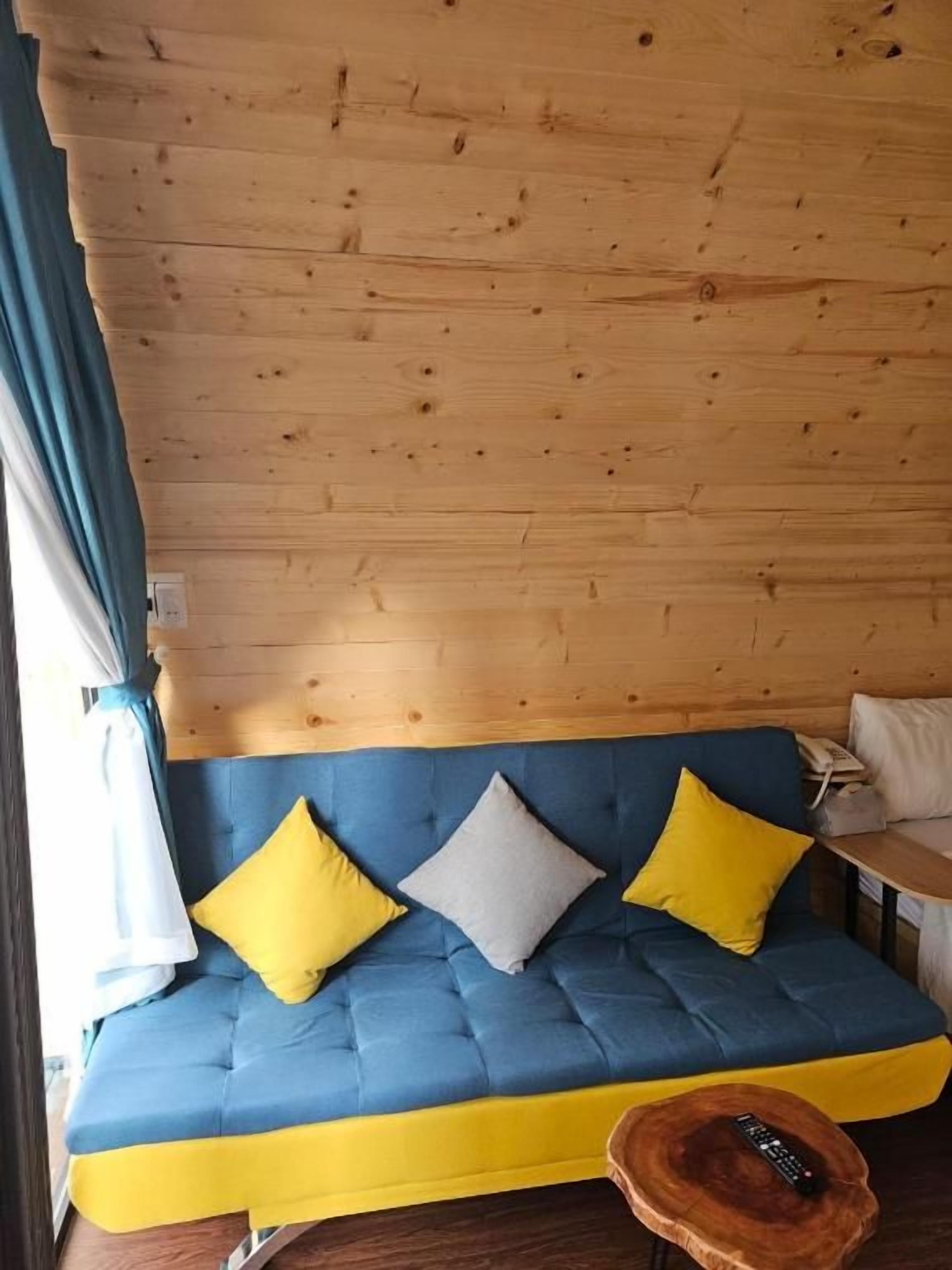 Techo de madera, sofá azul con cojines amarillos y grises, y una mesa redonda de madera con un control remoto.