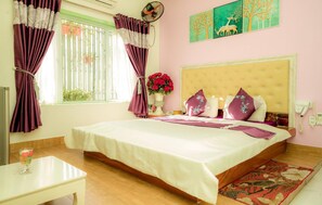 A bedroom with a bed, a bedside table, a window with curtains, a wall clock, a vase with flowers, and a painting on the wall. - Mio Hotel (Hai Phong)