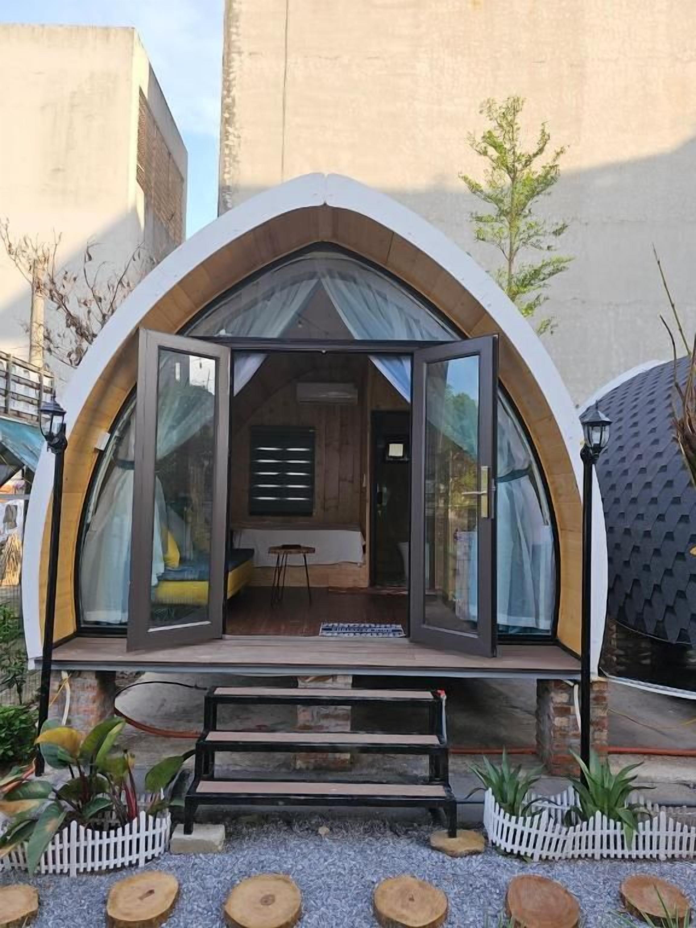 Estructura moderna, parcialmente abierta, con un banco y plantas en macetas al frente, y una pequeña mesa en su interior.