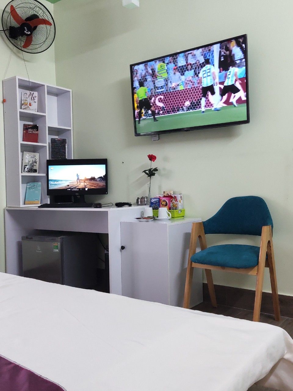 Habitació d'hotel amb un llit, un escriptori amb un ordinador, una televisió, un refrigerador petit, una estanteria, una cadira i una rosa sobre l'escriptori.