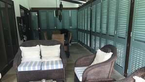 Et overdækket udendørs opholdsområde med fletmøbler, herunder en sofa og stole, samt et lille bord med kop og underkop.
