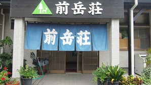 Un bâtiment sur lequel on peut lire l’inscription « 前島荘 » et qui est orné d’une banderole bleue portant le même texte. Des plantes en pot sont disposées devant l’entrée.