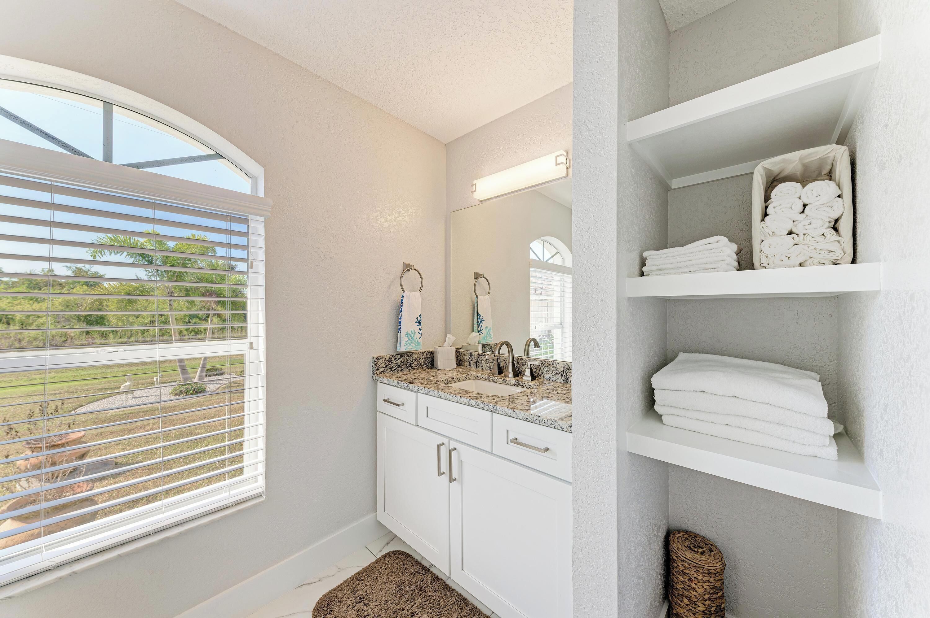 A bathroom with a large window, a sink with a granite countertop, and a mirror. There are shelves with towels and a basket on the floor.