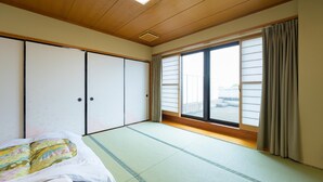 A room with tatami flooring, a bed with a colorful blanket, sliding doors, and a large window with curtains. - Hisai Green Hotel (Tsu)