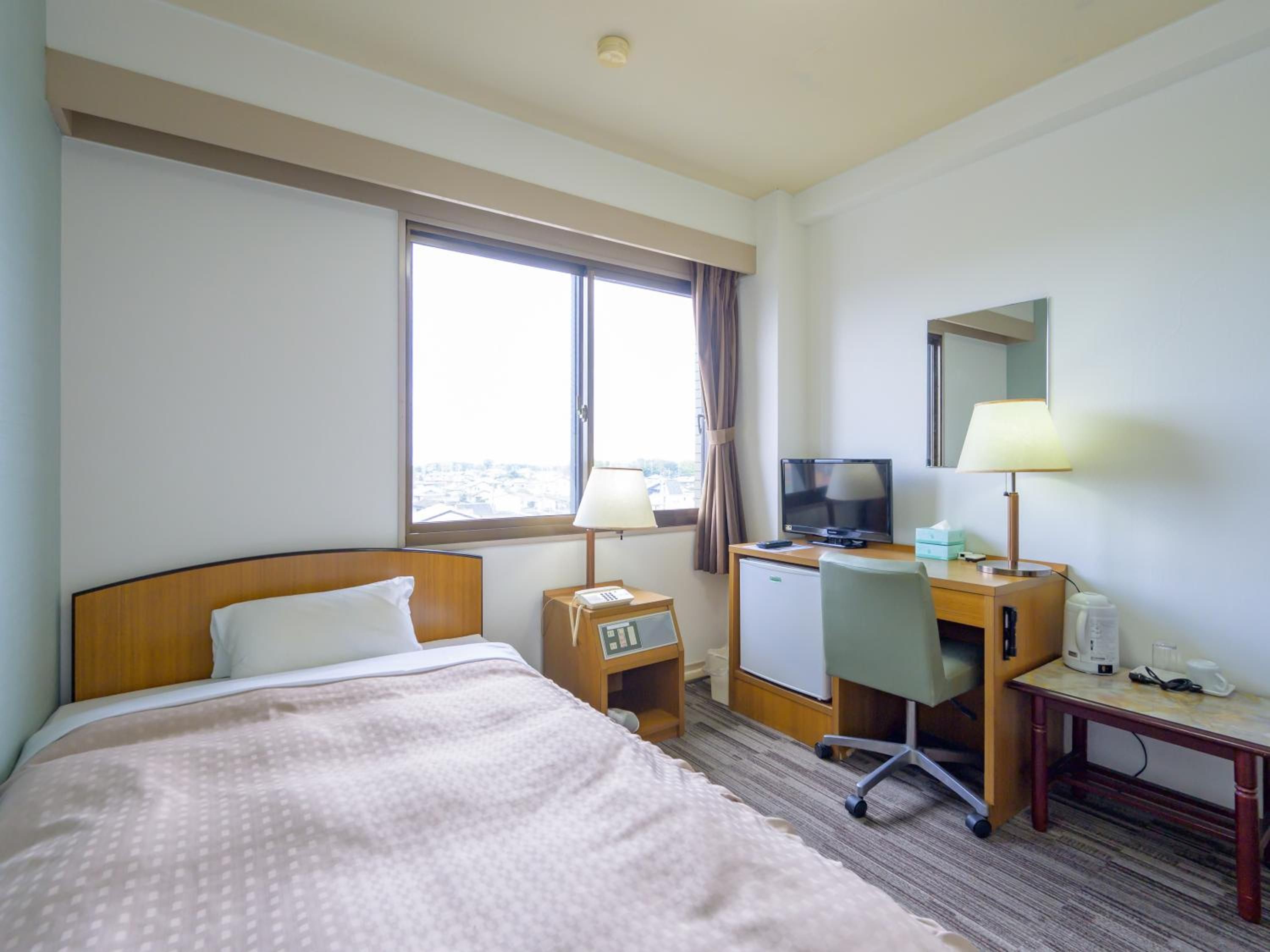 Habitación de hotel con cama, escritorio, silla, televisor, lámpara, reloj y ventana con vistas.