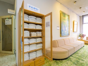 A hotel room with a wooden shelf filled with folded towels, a beige sofa, and a painting on the wall.