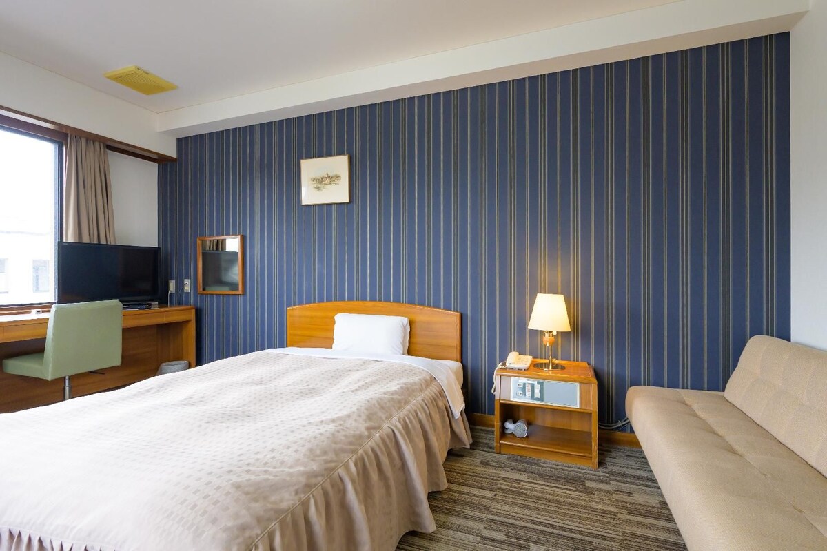 A hotel room with a bed, a desk with a chair, a TV, a bedside table with a lamp, and a sofa.