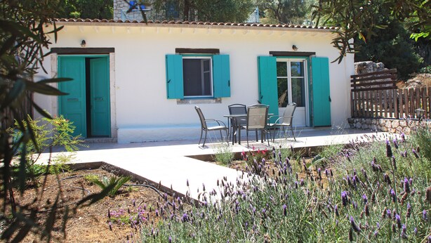 A small building with two doors and windows, a patio with chairs and a table, and a garden with purple flowers.