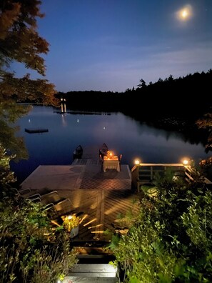 A wooden deck with a table and chairs overlooking a calm lake at night. - 5 star cottage, private, 62 acres, lakefront, hot tub, boat, sauna, pet-friendly (Shelburne)