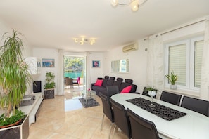 A modern living room with a dining area, a sofa with red cushions, a coffee table, and a TV. There are potted plants and a large window with blinds.