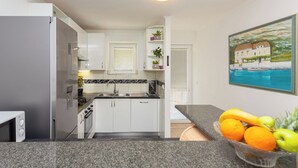Une cuisine avec des meubles blancs, un four encastré, un four à micro-ondes et un panier de fruits posé sur le plan de travail.