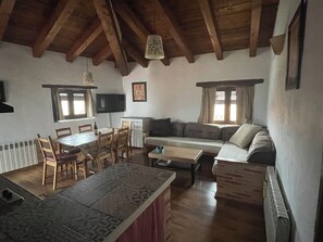 A cozy living room with a wooden ceiling, a dining table with chairs, a sofa, and a TV mounted on the wall.