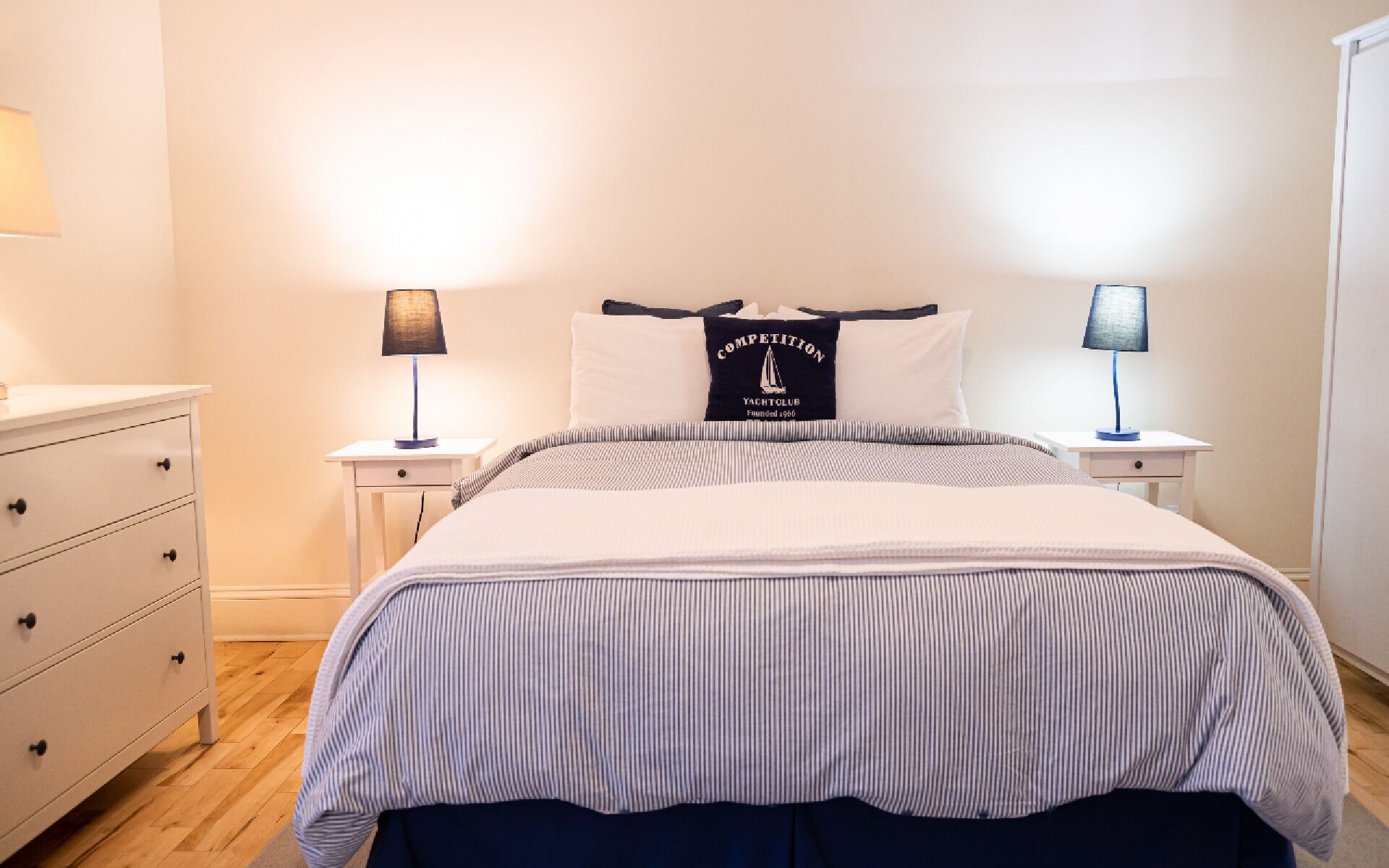 A neatly made bed with striped bedding, two bedside tables with lamps, a dresser, and a pillow with a logo.
