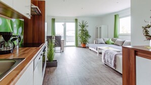 Una cocina moderna con encimera de madera y estufa integrada. Hay una zona de comedor con mesa y sillas blancas, y un ventanal con cortinas verdes. Se ve una planta en maceta en primer plano.