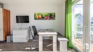 Una habitación moderna con televisión de pantalla plana, un mueble de madera, un escritorio con silla y un banco. Hay un cuadro en la pared y una ventana con cortinas verdes.