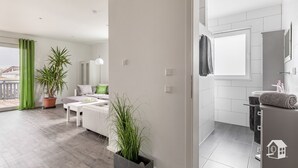 Un apartamento moderno con balcón, sala de estar con sofá y mesa de centro, zona de cocina y baño con lavamanos y perchero.