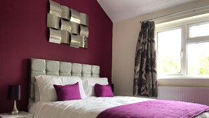 Ein Schlafzimmer mit einem karomustertem Kopfteil, weißer Bettwäsche, violetten Kissen, einer violetten Tagesdecke, einer Nachttischlampe und einem Fenster mit Vorhängen.