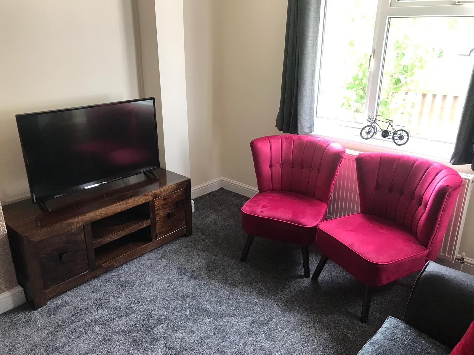 A living room with a flat-screen TV on a wooden stand, two red velvet armchairs, and a window with a bicycle outside.