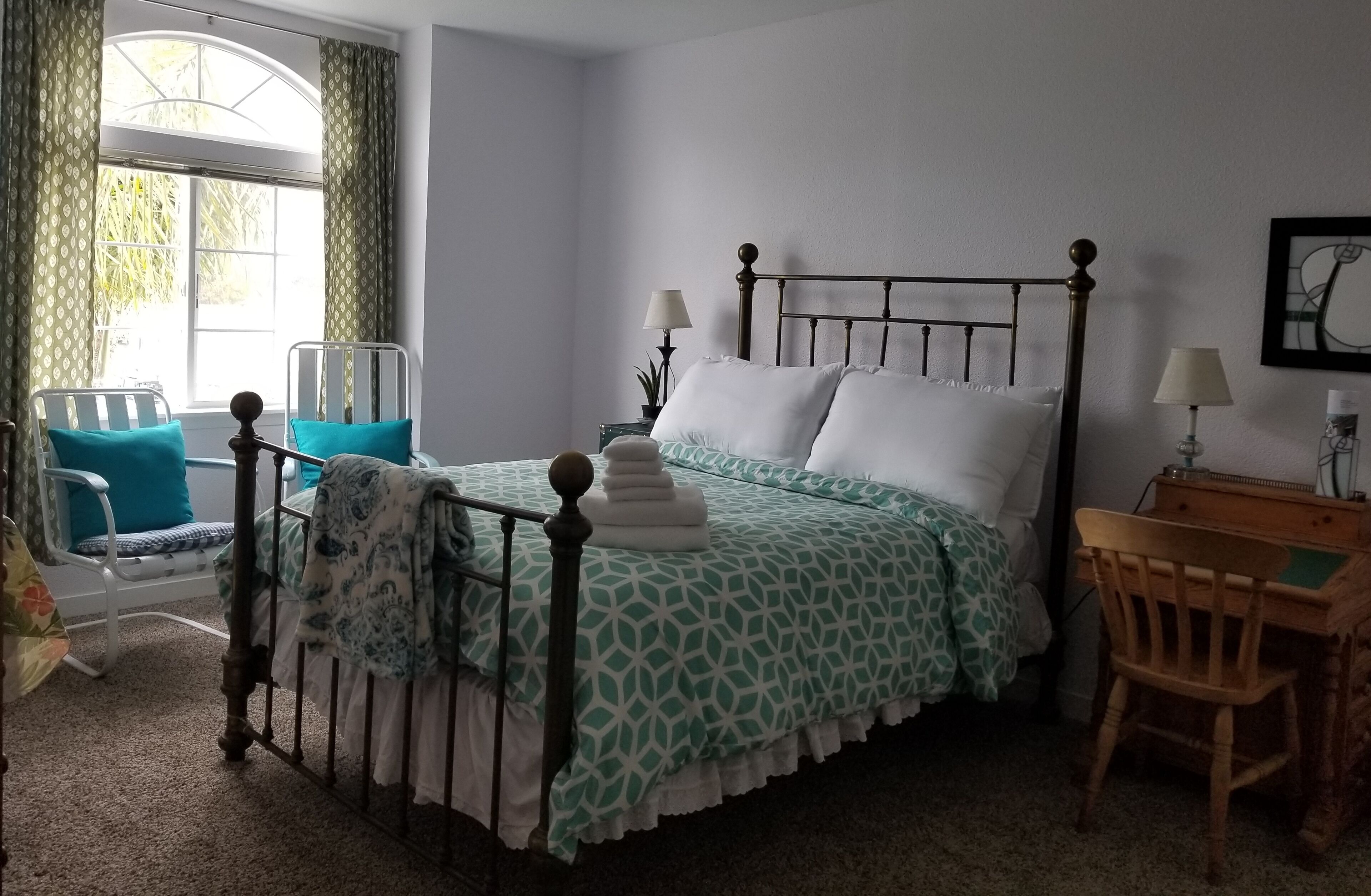 A bedroom with a four-poster bed, a desk with a chair, a window with curtains, and a bedside table with a lamp.