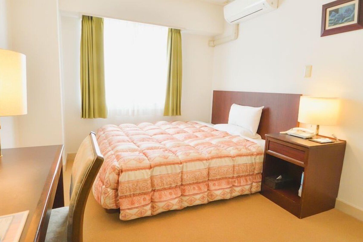 A hotel room with a bed, a nightstand, a desk, and a window with curtains.