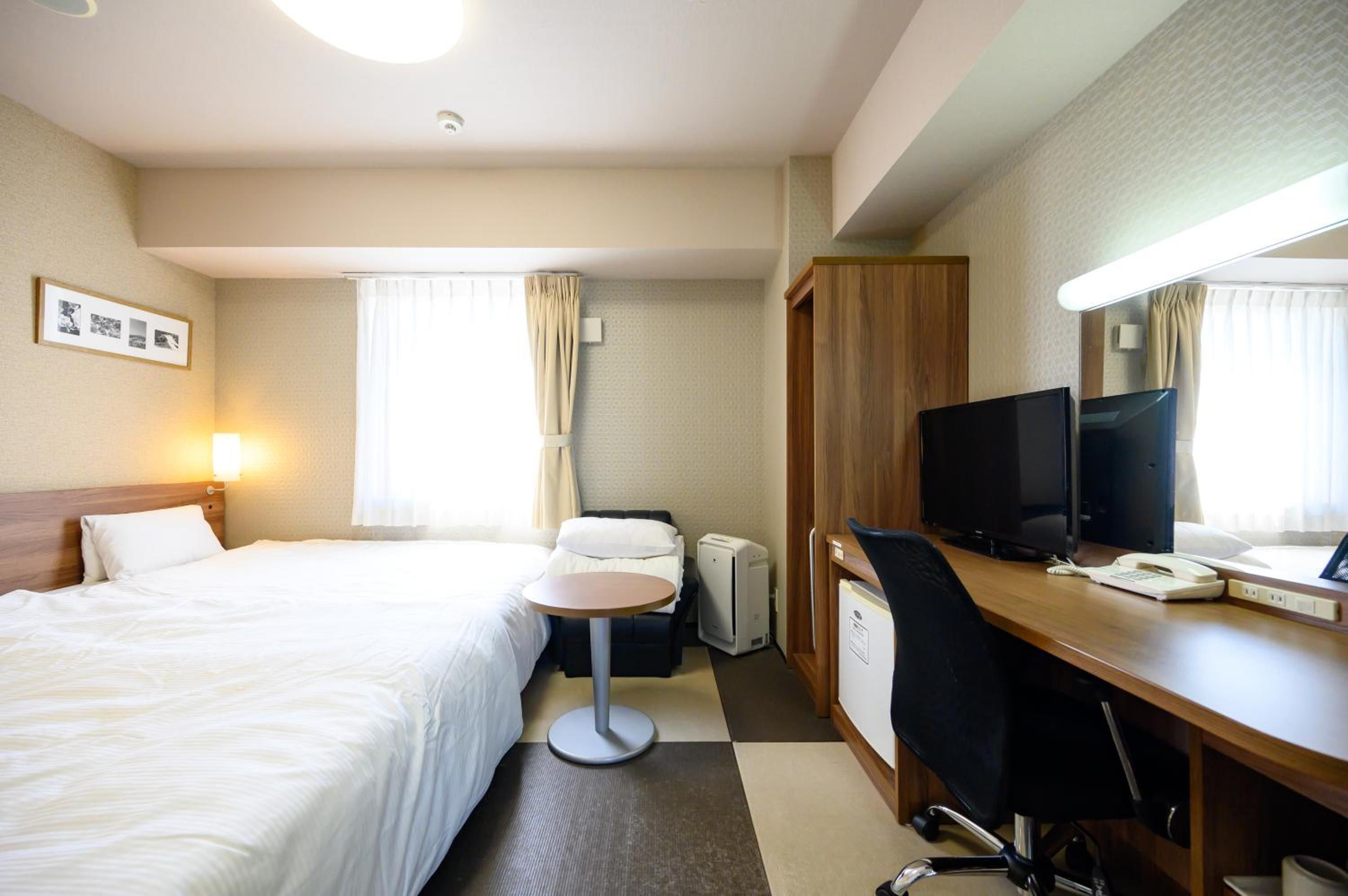 A hotel room with a bed, a desk with a computer, a TV, and a small table.