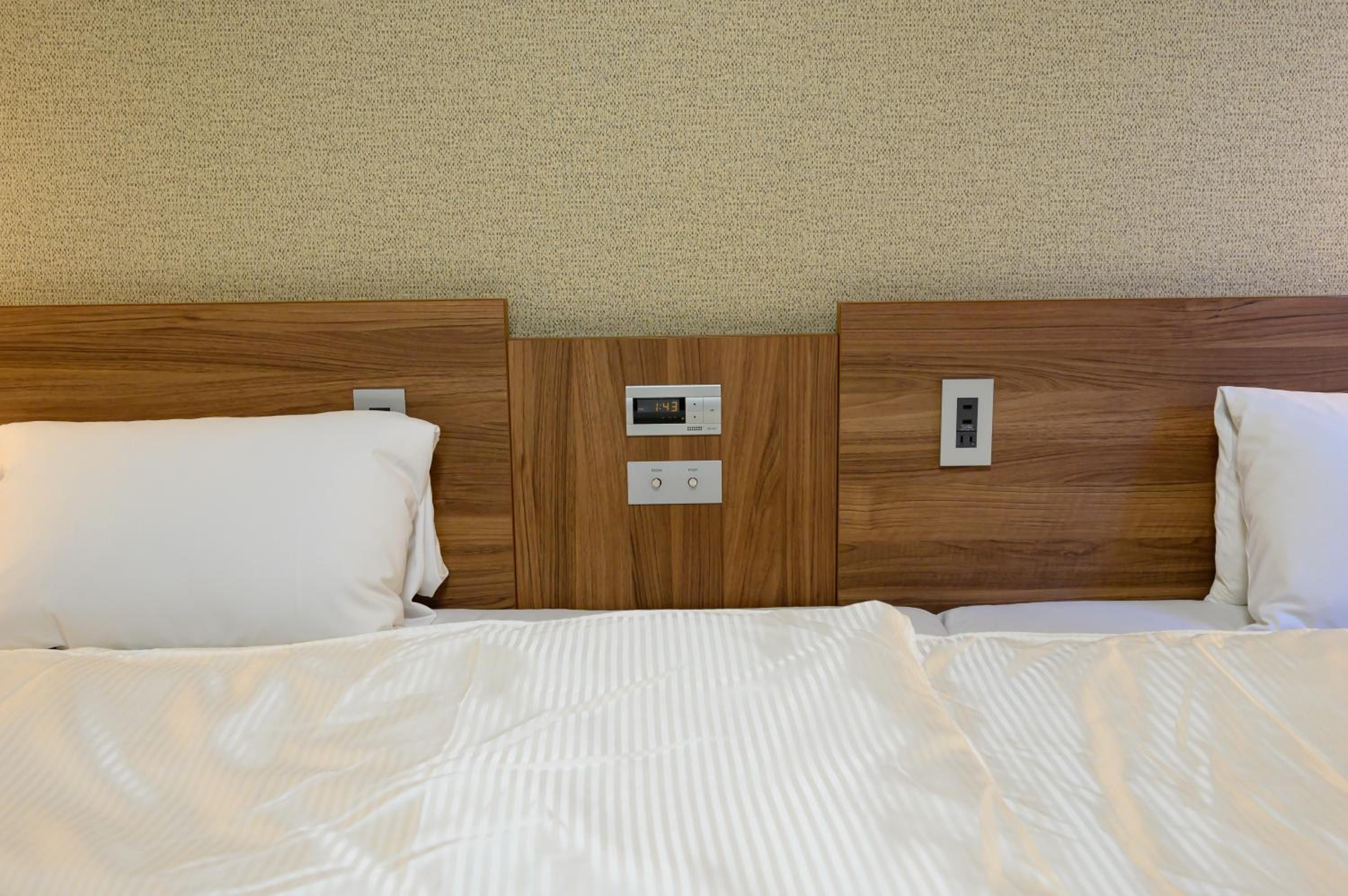 A hotel room with a wooden headboard, two white pillows, a bed with white bedding, and a digital clock displaying the time.