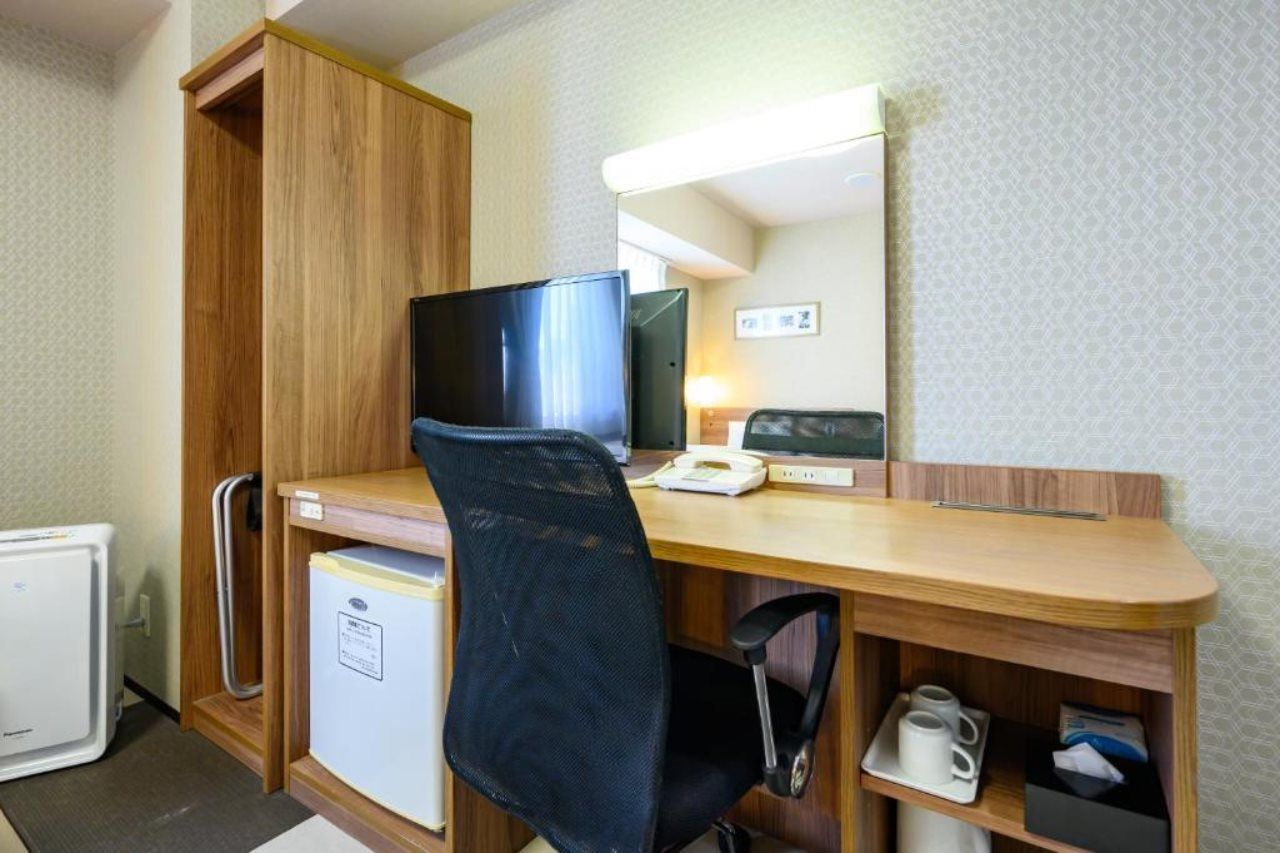 A compact hotel room with a wooden desk, a television, a mini-fridge, a chair, and a small shelf with a cup.