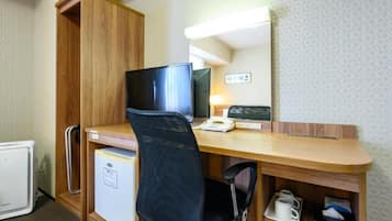 A compact hotel room with a wooden desk, a television, a mini-fridge, a chair, and a small shelf with a cup.