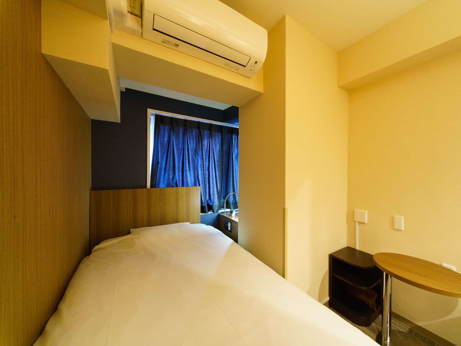 A hotel room with a bed, a small table, a desk with a chair, an air conditioning unit, and a window with curtains.