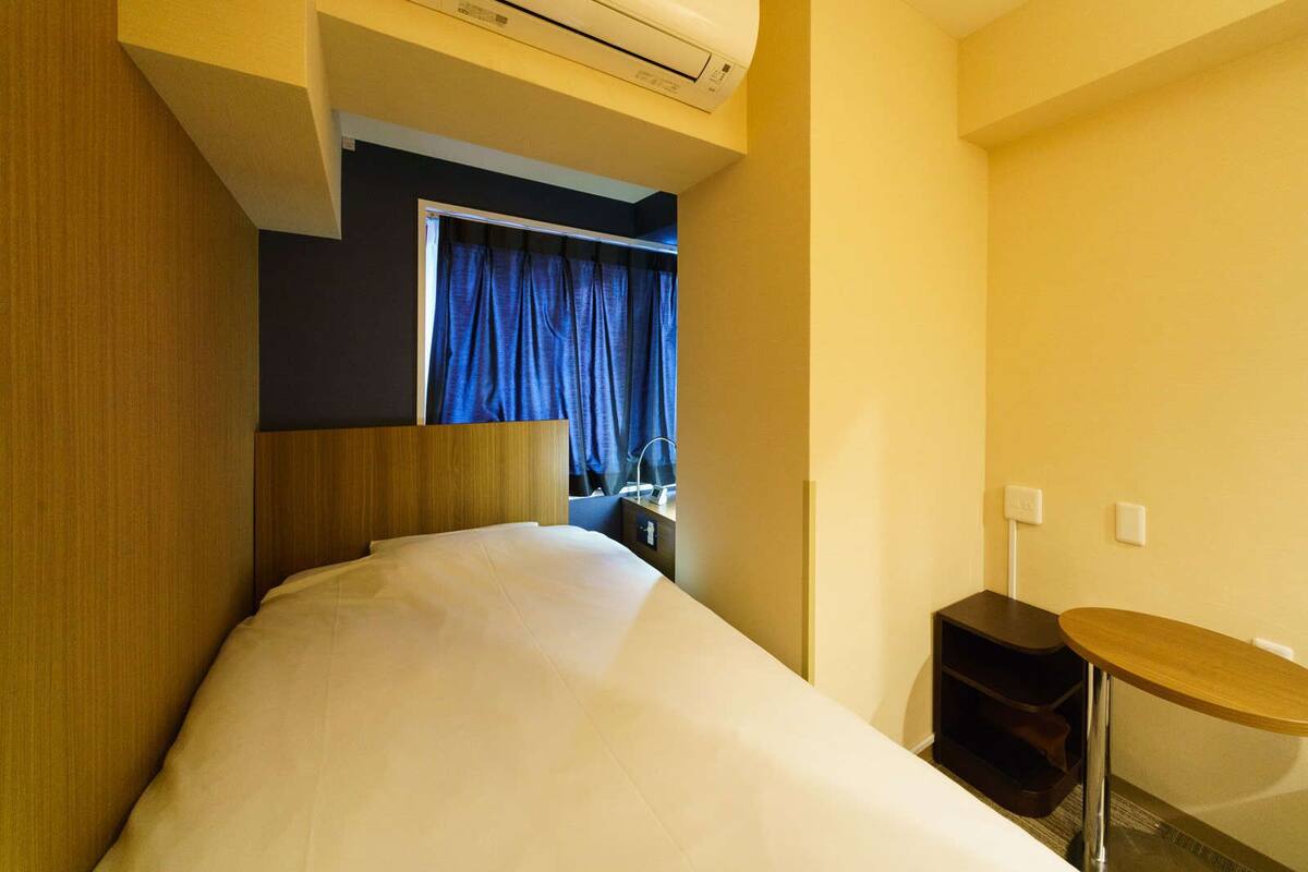 Habitación de hotel con cama, una mesita, un escritorio con silla, aire acondicionado y ventana con cortinas.