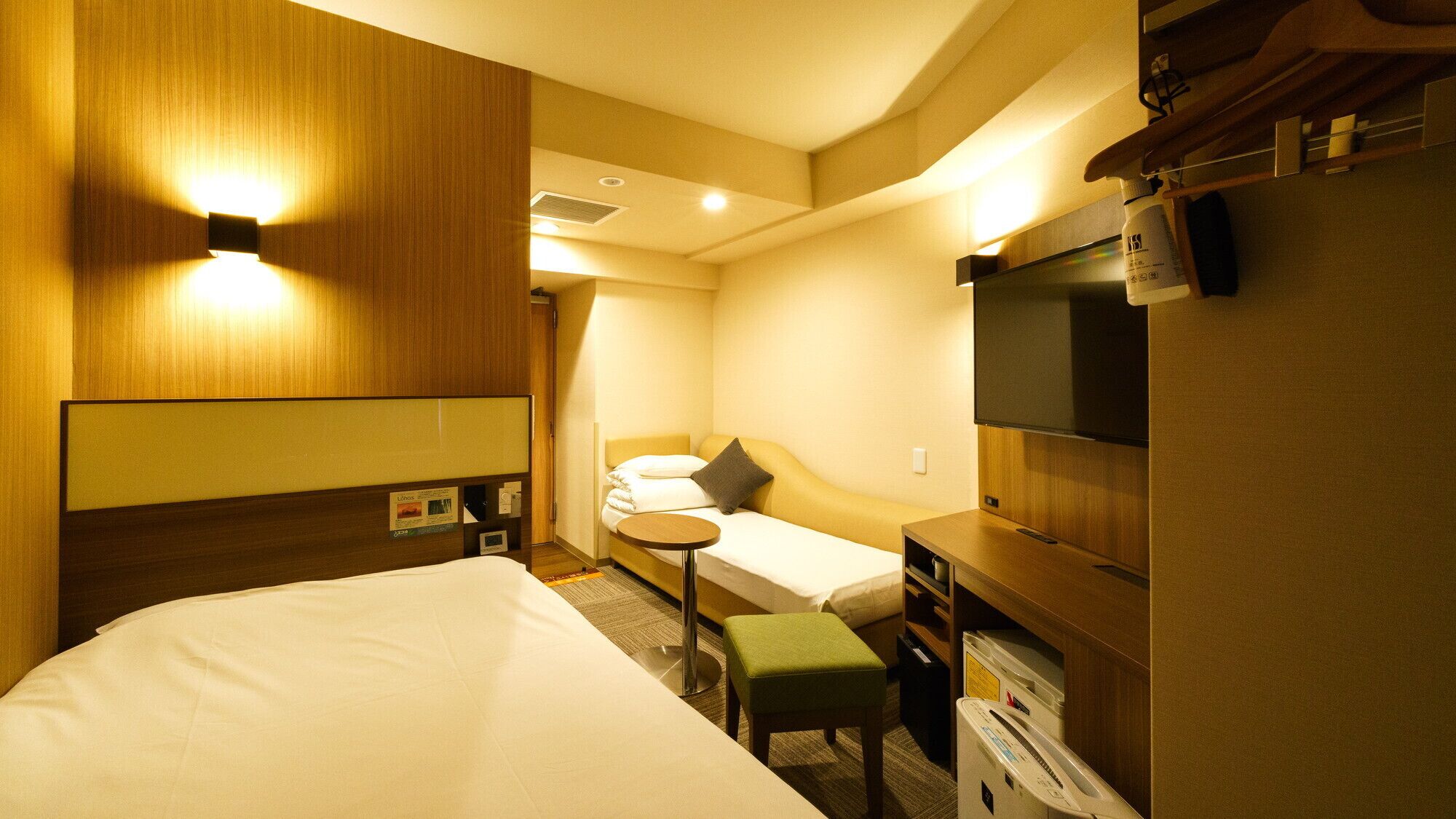 A hotel room with a large bed, a smaller bed, a desk with a chair, a TV, and a minibar.