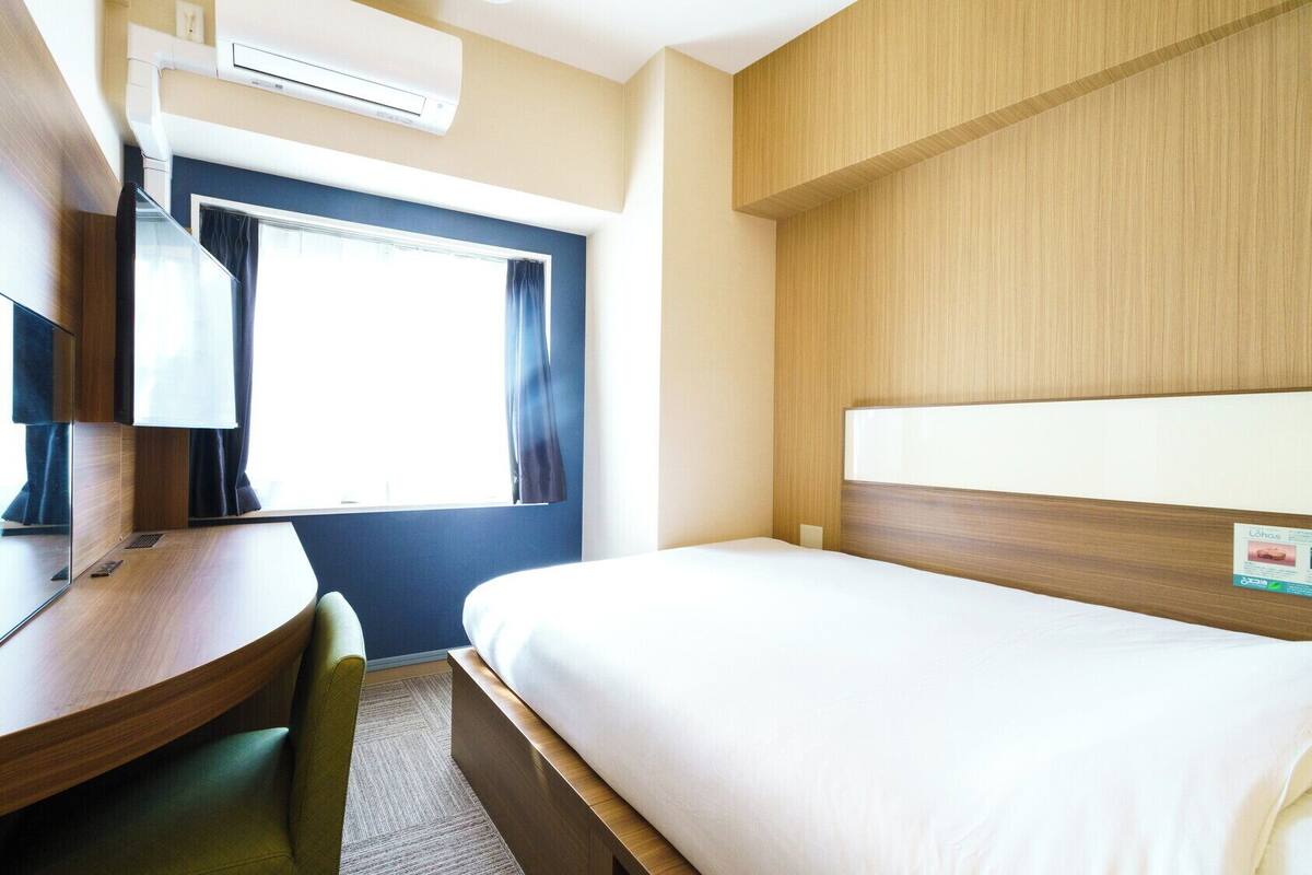 Habitación de hotel con cama, escritorio, silla, ventana con cortinas y aire acondicionado.