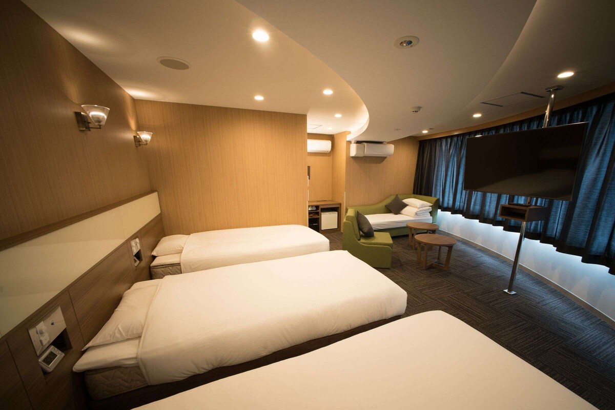 Ein Hotelzimmer mit zwei Einzelbetten, einem Schlafsofa, einem kleinen Tisch und einem an der Wand montierten Fernseher.